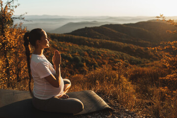Naklejka na ściany i meble Spiritual and emotional concept of harmony with nature in maternity time. Pregnant woman praying alone outdoors on hill at sunset. Amazing autumn mountain view.