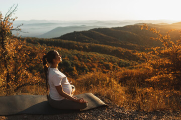 Naklejka na ściany i meble Pregnant woman in lotus position do yoga alone outdoors. Amazing autumn mountain view at sunset. Spiritual maternity concept, natural harmony.