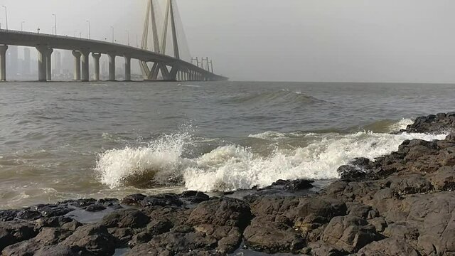 Small waves crashes on rocks at Bandra fort with view of Bandra-Worli Sea Link bridge in the background in Mumbai, slow motion shot.