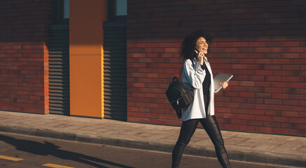Fototapeta premium Walking caucasian woman with curly hair is talking on phone and holding a laptop