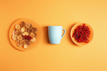 Obraz na płótnie Canvas Homemade mini pancakes in an orange plate on an orange background, next to it is a blue ceramic mug and there is a plate with red currants, flat lay, healthy breakfast concept, knolling.