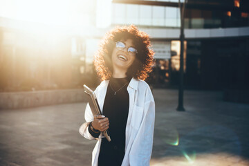 Curly haired businesswoman wearing eyeglasses and smiling at camera is posing in a sunny day with a...