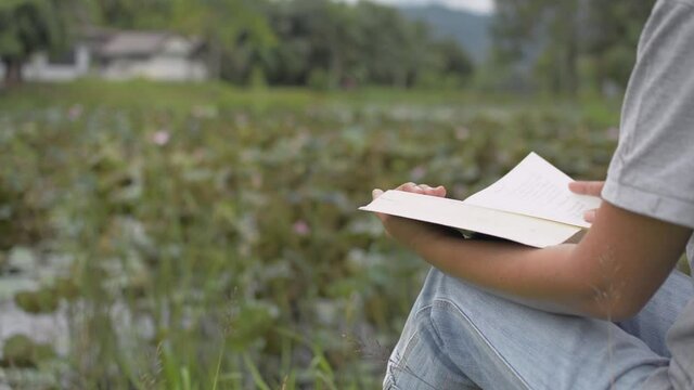 Close up asian woman in casual dress is sitting on the grass near natural pond with lotus and reading a novel happily. Hand turning pages of a book. Education.