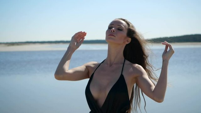 The statuesque, young lady in a light summer dress is black with a large neckline and open back straightens her hair and hugs herself on the beach against the backdrop of the lake