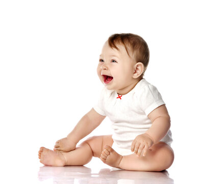 Laughing loud adorable little baby infant toddler in white cotton bodysuit sits on floor touching legs, looking aside