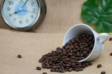 Roasted coffee beans In a white coffee cup on its side and a lot of coffee beans spread on the floor and an alarm clock placed behind the picture and the bottom surface is a brown fabric.