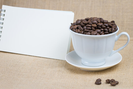 Roasted coffee beans In a white coffee cup and with a blank notebook and the bottom surface is a brown fabric and a few coffee beans scattered on the floor and the picture has a blank space