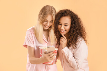 Beautiful young women in pajamas, with popcorn and mobile phone on color background