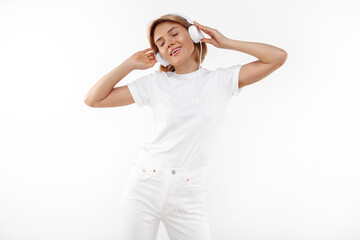 Modern young blonde woman in casual white outfit listening music in headphones over white background.