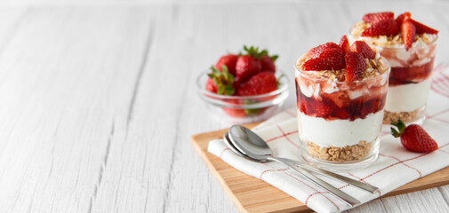 Homemade layered dessert with fresh strawberries, cream cheese or yogurt, granola and strawberry jam in glasses on white wood background. Healthy organic breakfast or snack concept. Selective focus.