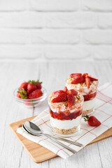 Homemade layered dessert with fresh strawberries, cream cheese or yogurt, granola and strawberry jam in glasses on white wood background. Healthy organic breakfast or snack concept. Selective focus.