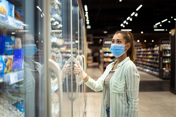 Woman with protective mask and gloves opening freezer in supermarket during COVID-19 pandemic or corona virus. Protect yourself against highly contagious coronavirus.