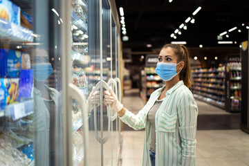 Fototapeta premium Woman with protective mask and gloves opening freezer in supermarket during COVID-19 pandemic or corona virus. Protect yourself against highly contagious coronavirus.