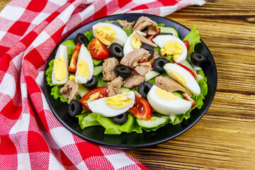 Tasty tuna salad with lettuce, black olives, eggs and fresh vegetables on wooden table