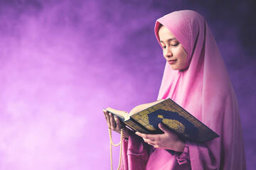 Asian Muslim woman in a veil holding prayer beads and reading the Quran with a colored background