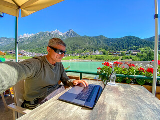 Relaxed man working with laptop in front of a mountain lake. Business and tourism concept