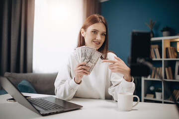 Competent female blogger in white sweater recording video on smartphone while sitting at table and holding money in dollars banknotes. Concept of technology, people and videoblog