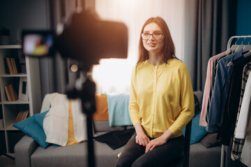 Fototapeta na wymiar Cheerful young woman in eyeglasses and casual outfit sitting on chair at bright room and shooting new video for blog. Young brunette talking to her followers in social networks.