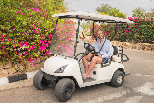 Friendly Man Takes Off His Hat While Sitting In A Golf Cart. A Cheerful Tourist Is Sitting Behind The Wheel Of An Electric Car Near Flowering Bushes.