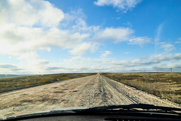 View from car windscreen with stripe relief to dirt country road, tundra and blue sky with white clouds in norht region at a sunny day