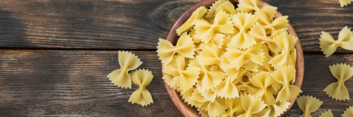 Raw Farfalle macaroni in a wooden bowl on a brown wooden table. Farfalle macaroni close-up. Banner with space for text