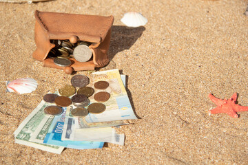 Fototapeta na wymiar Paper banknotes lie on the sea sand under the coins of different countries. Nearby is a purse made of clay filled with coins. The concept of saving finances for vacation, rest at the sea