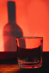 Low glass for scotch or whiskey and shadow of bottle. Container for alcoholic beverages. Red lighting indoors.