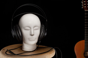 Ceramiczna biała głowa z nałożonymi studyjnymi słuchawkami, stojąca na drewnianym taborecie, w tle fragment gitary klasycznej. - obrazy, fototapety, plakaty