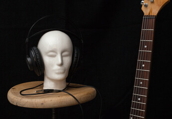 Ceramiczna biała głowa z nałożonymi studyjnymi słuchawkami, stojąca na drewnianym taborecie, z fragmentem gitary elektrycznej. - obrazy, fototapety, plakaty