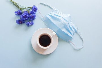 Medical protective face mask from coronavirus Covid-19, cup of coffee and blue flowers on a pastel blue background. Biological concept of life in a new reality. Top view, flat lay lifestyle.