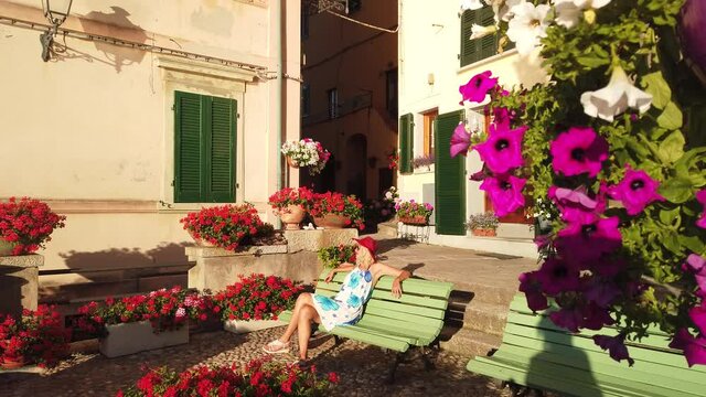 Woman sitting on bench in Marciana Marina, flowery old district Borgo al Cotone. Tourist travel in Italy. Marciana Marina, Elba island. Blooms of red geraniums on foreground.