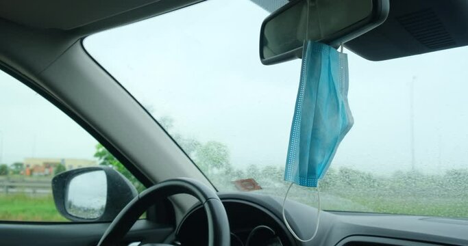 Medical mask hangs from car mirror, on a rainy day. Essential personal protection during Coronavirus (Covid-19) pandemic.