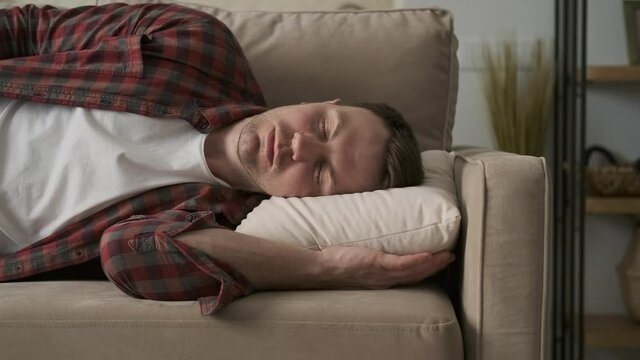 Tired man in red shirt sleeping on couch. Portrait of handsome guy napping on sofa at home. Young guy relaxing in 4K, UHD