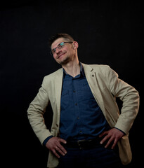 smiling man in beige classical jacket and glasses bends head with hands on hips against black background close view