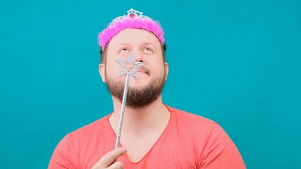 funny bearded freaky man in a pink T-shirt with a deadema on his head is dreaming with a magic wand...