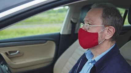 Old grey-haired business man wears protective face mask in prevention of coronavirus infection and driving his car through city. Pandemic and healthcare. Public safety. Transportation. - Powered by Adobe