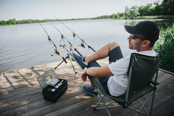 Obraz premium Young fisherman fishing on lake or river. Side view of adult guy sitting in folding chair alone at river or lake shore. Man fishing by kepping three rods beside him in water.