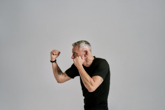 Middle aged athletic man in black sportswear boxing, practicing punches while exercising in studio over grey background
