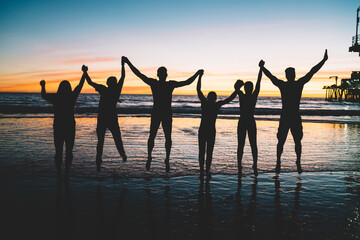 Fototapeta na wymiar Happy young male and female persons holding each other hands standing on beach at scenery sunset, silhouette group of people enjoying summer vacations together and jumping in air, Friendship concept