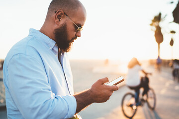 Concentrated hipster guy read news from browsed website on smartphone spending free time in city outdoors, handsome male using modern cellular and 4G connection for chatting in social networks online