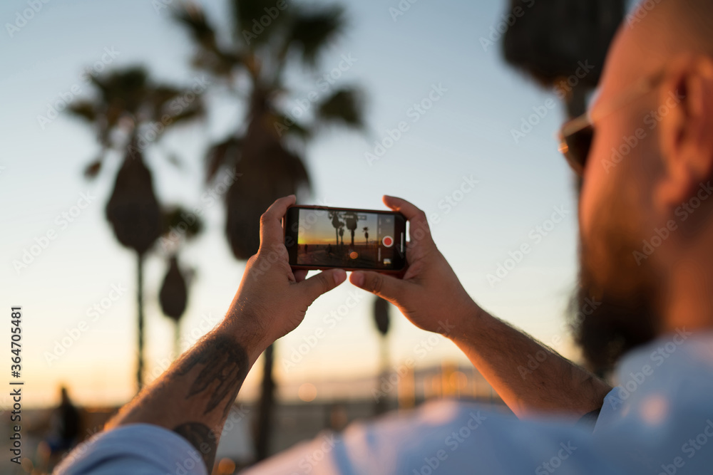 Poster back view of young man using smartphone camera for taking pictures of sunset and share in social net