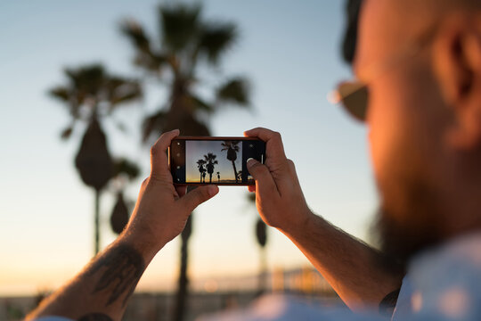 Close up view of male tourist shooting video of beautiful scenery sunset using smartphone camera, cropped view of hipster guy holding modern telephone for taking photos of exotic palm trees in evening