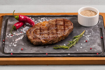 A RIBEYE STEAK is on a black basalt Board on a wooden background with demiglas sauce. Perfectly crusted with foaming warm butter infused with smashed garlic cloves and fresh thyme + rosemary sprigs.