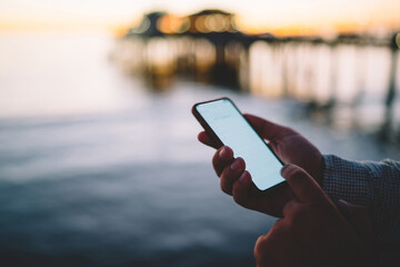 Cropped image of man checking notification on smartphone with mock up screen near sea in evening, male hands holding modern mobile phone sending message via good 4G connection to internet in dusk