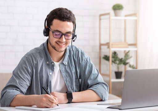 Student at distance learning at home. Guy in glasses and headset makes notes in notebook, near laptop
