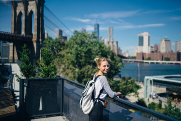 Positive young woman enjoying beautiful urban scenery near bridge over river during trip to...