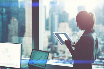 Female employee work on digital tablet standing at office window with skyscrapers view.Silhouette...