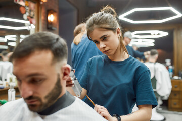 Focused professional barber girl working with hair clipper, making modern haircut for a young handsome man visiting barbershop
