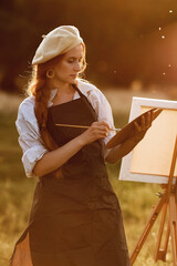  a girl artist with long red hair draws on an easel with a brush against the background of the sunset