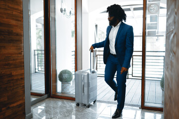 Black man in formal suit with packed suitcase entering hotel door